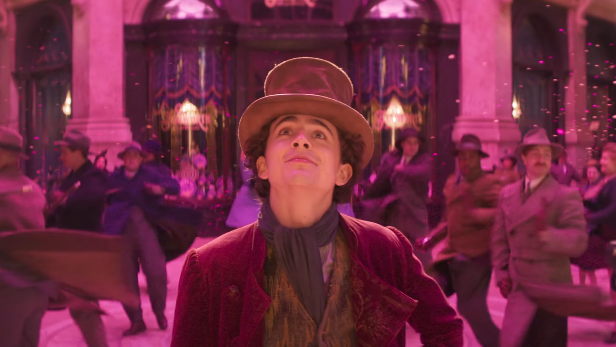 Timothee Chalamet w roli Willy'ego Wonki, ma cylinder, różowy frak, patrzy rozmarzony na górę, w tle widać tańczący tłum
